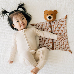 Child laying on a bed wearing a Heather Oatmeal zippy next to a Bowie the Bear lovey