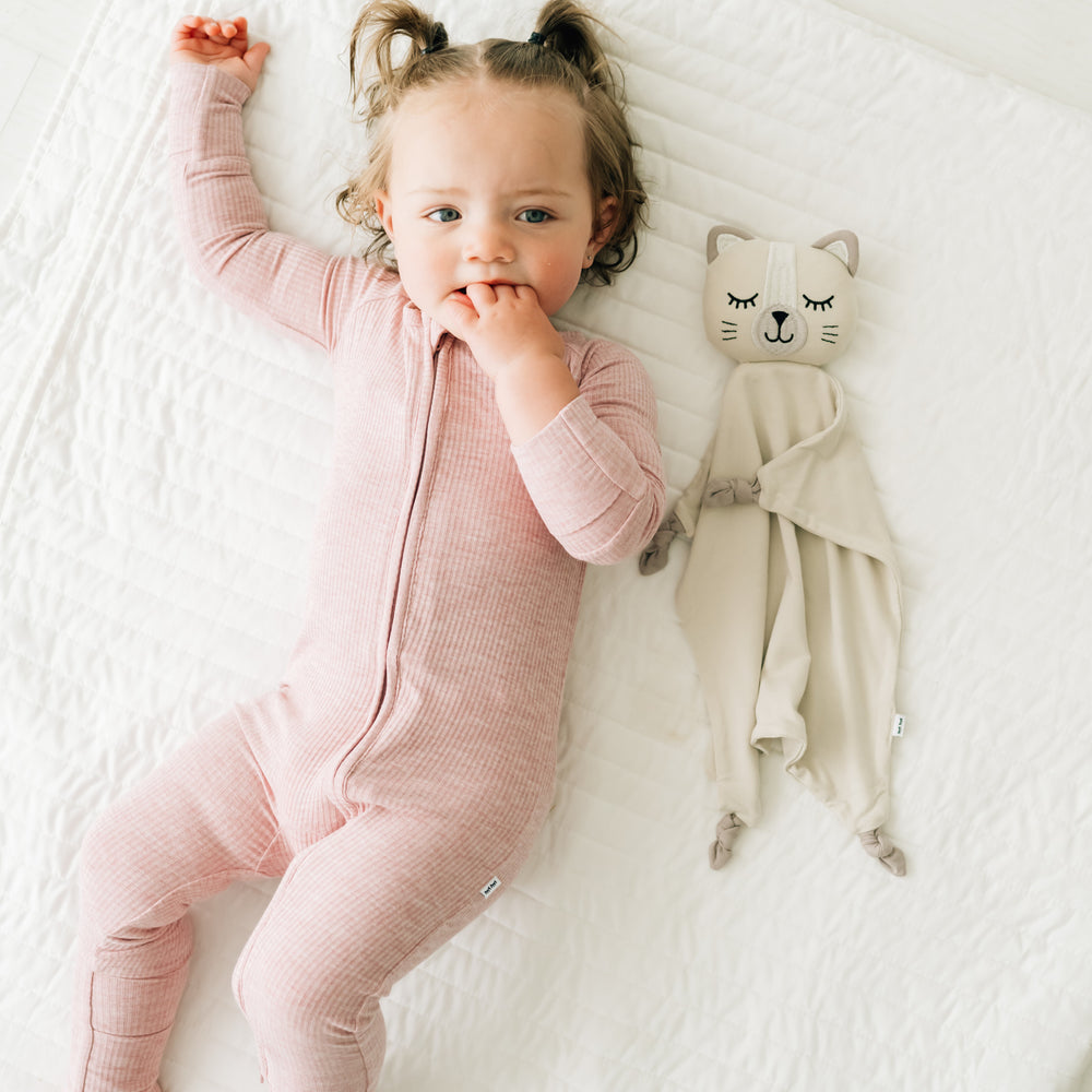 Child laying on a bed wearing a Heather Mauve zippy next to her Clementine the Cat lovey