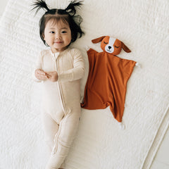 Child laying on a bed wearing a Heather Oatmeal zippy next to her Parker the Puppy lovey