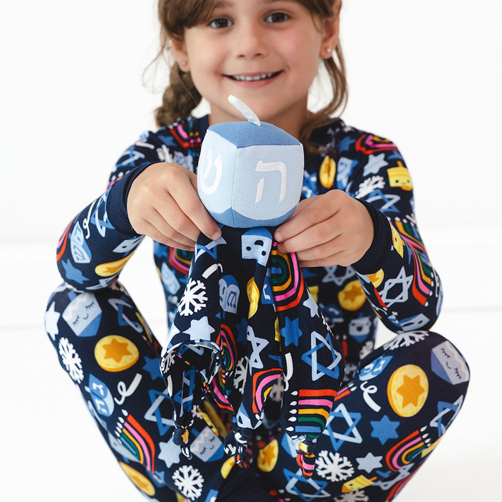 Child sitting on the ground holding a Spin the Dreidel Sleepyhead Lovey and wearing Playful Dreidel pajamas