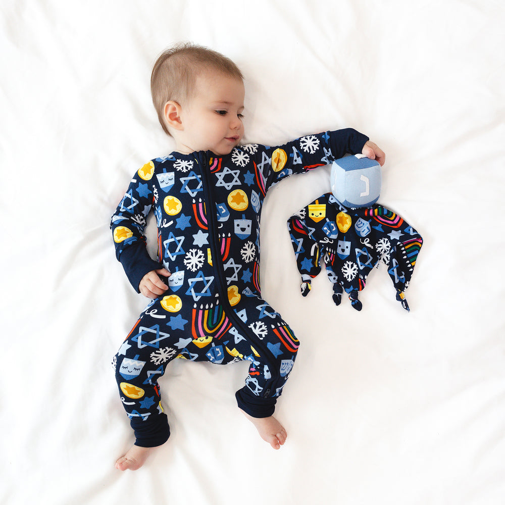 Child laying on a blanket next to a Spin the Dreidel Sleepyhead Lovey and wearing a Playful Dreidels Zippy