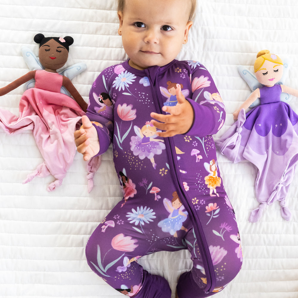 Top view image of child laying down next to Fiona the Purple Fairy Sleepyhead Lovey & Flora the Pink Fairy Sleepyhead Love, while wearing the Purple Fairy Zippy