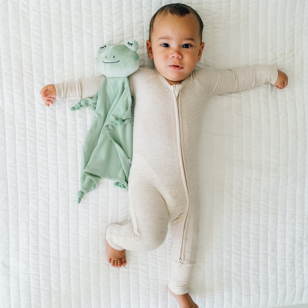 Child laying on a bed wearing a Heather Oatmeal zippy with a Fredrik the Frog lovey