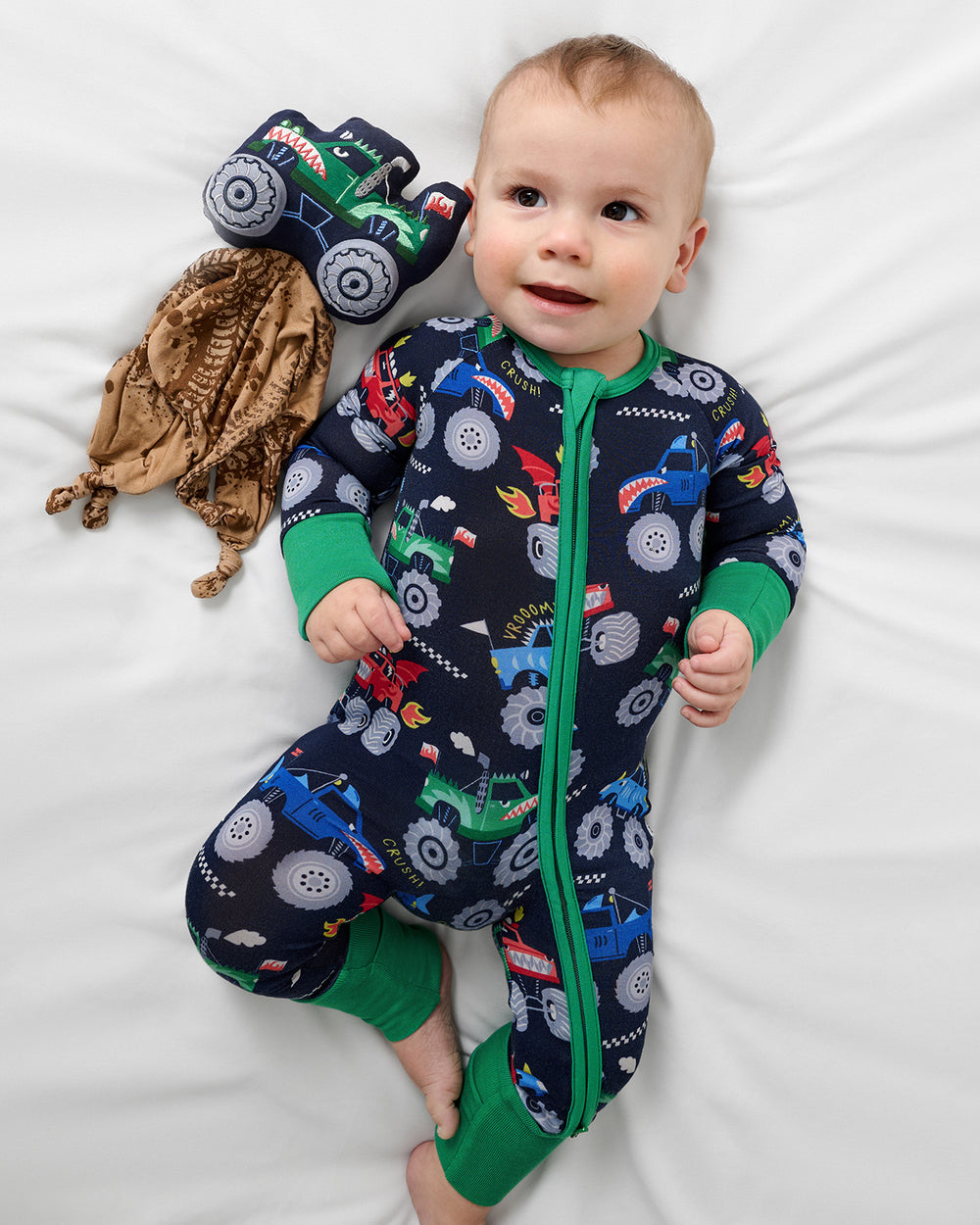 Aerial view of baby laying next to the Max the Monster Truck Sleepyhead Lovey in a matching footie pajama