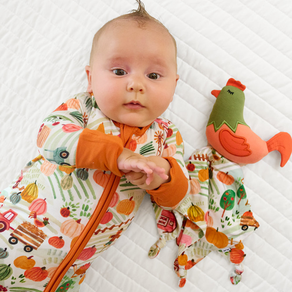 Child laying on a bed wearing a Happy Harvest zippy next to a Rocky the Rooster sleepyhead lovey
