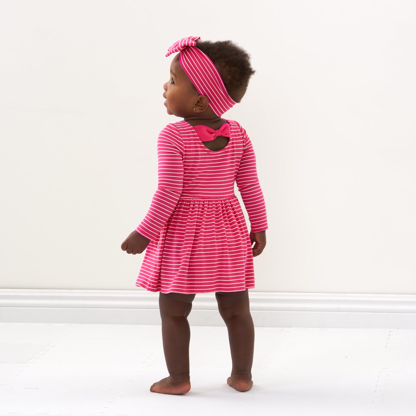 Back view image of a child wearing a Pink Punch Stripes bow back skater dress with bodysuit and matching Luxe Bow Headband