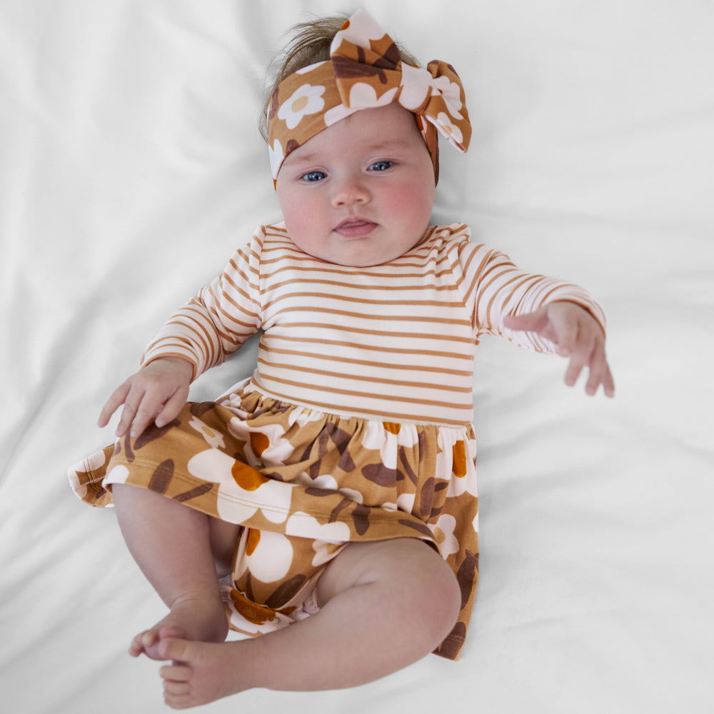 Infant lying down wearing a Daisy Daydream twirl dress with bodysuit paired with a tan floral bow headband