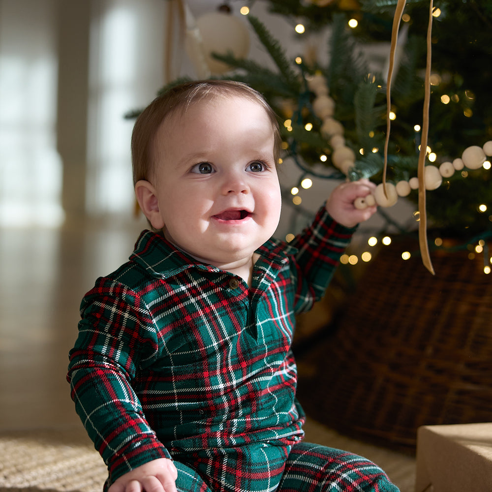 Close up image of a child sitting in front of a Christmas tree wearing an Evergreen Plaid Polo Romper