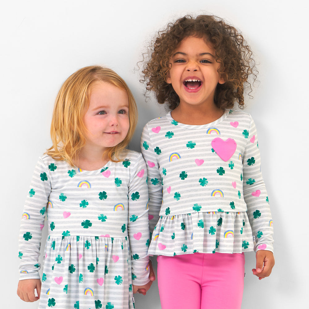 Click to see full screen - Two children posing together wearing coordinating Lucky in Love play styles. One child is wearing a Lucky in Love pocket peplum tee paired with sweet pink leggings. Second child is wearing a Lucky in Love skater dress