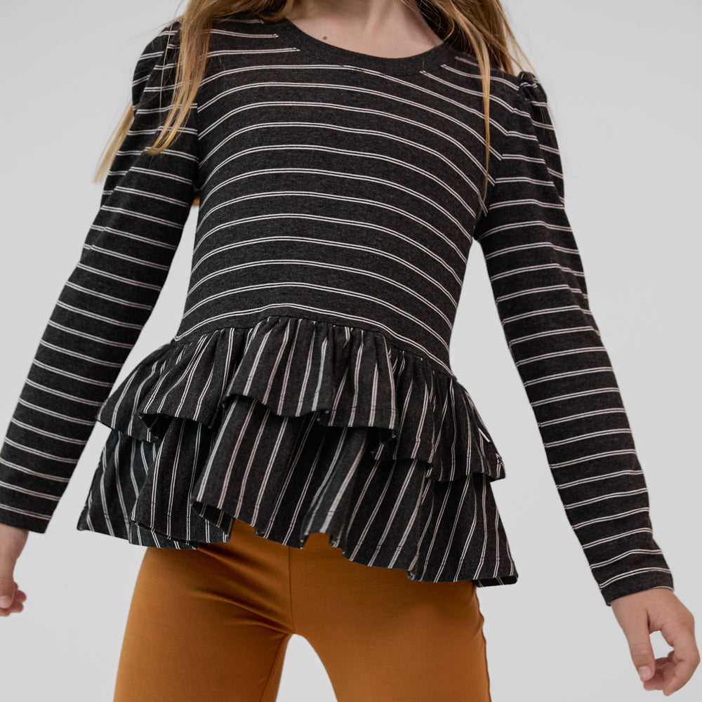 Close-up view of a child wearing a Heather Black Stripes long sleeve tiered peplum top paired with golden brown leggings