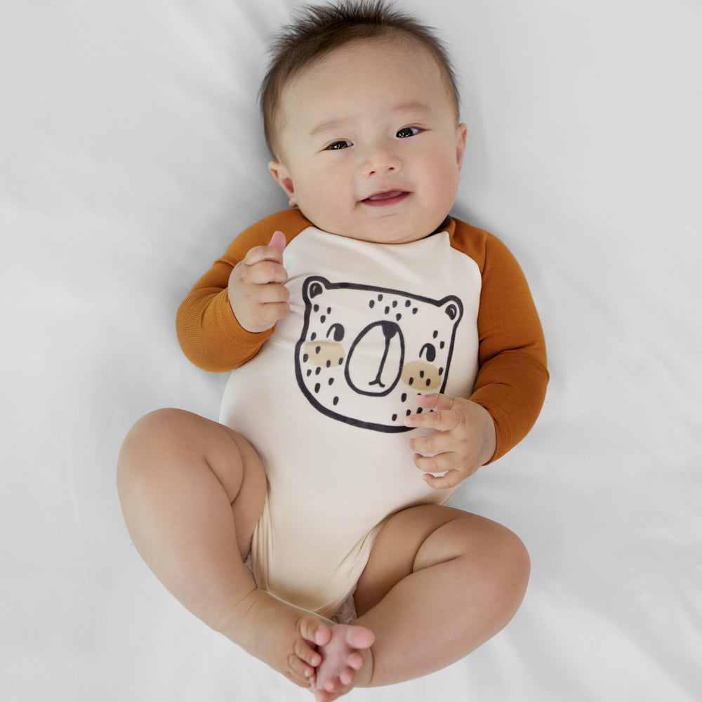 Child lying on a blanket wearing an Arctic Raglan baby bodysuit featuring tan long sleeves and a black bear illustration