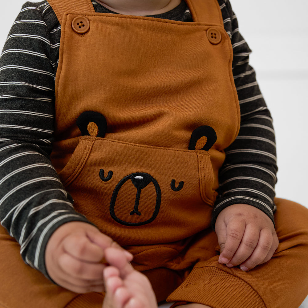 Close-up view of a child wearing a Golden Brown graphic overall paired with a black striped long sleeve tee