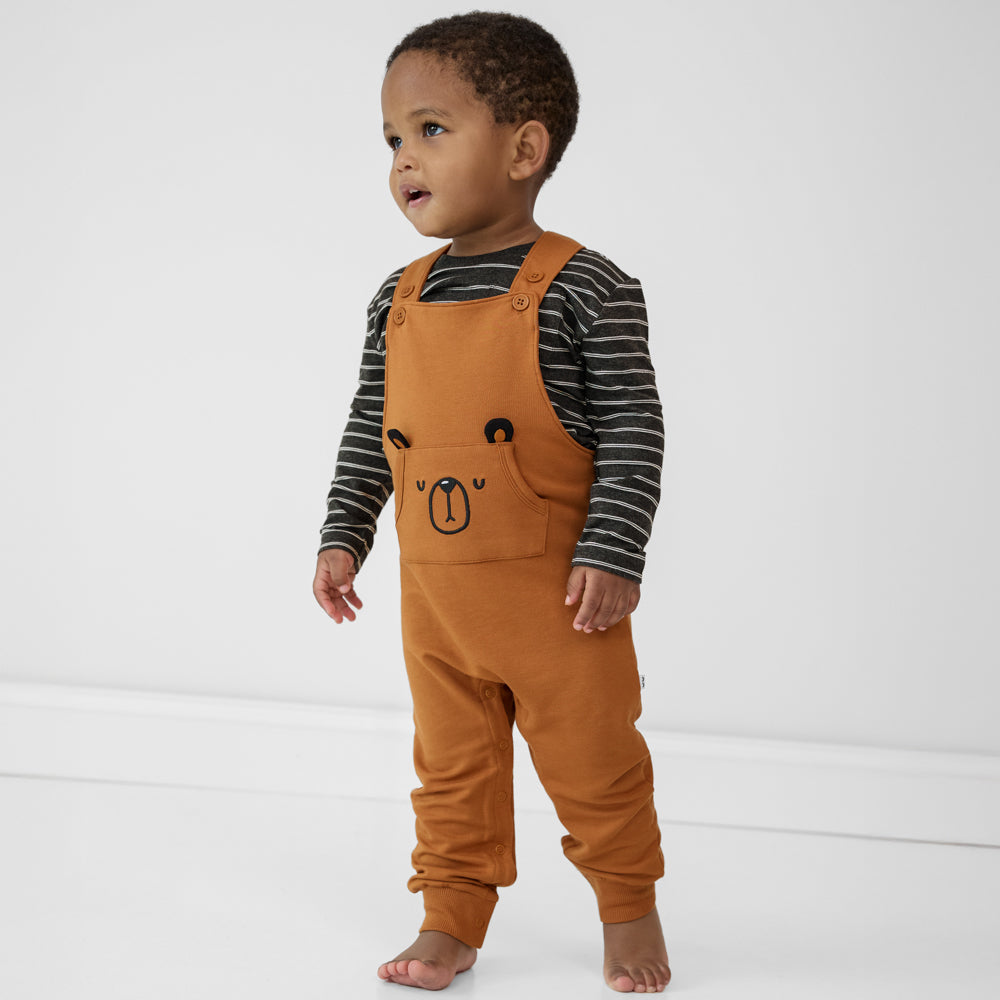 Child wearing a Golden Brown graphic overall paired with a black striped long sleeve tee.