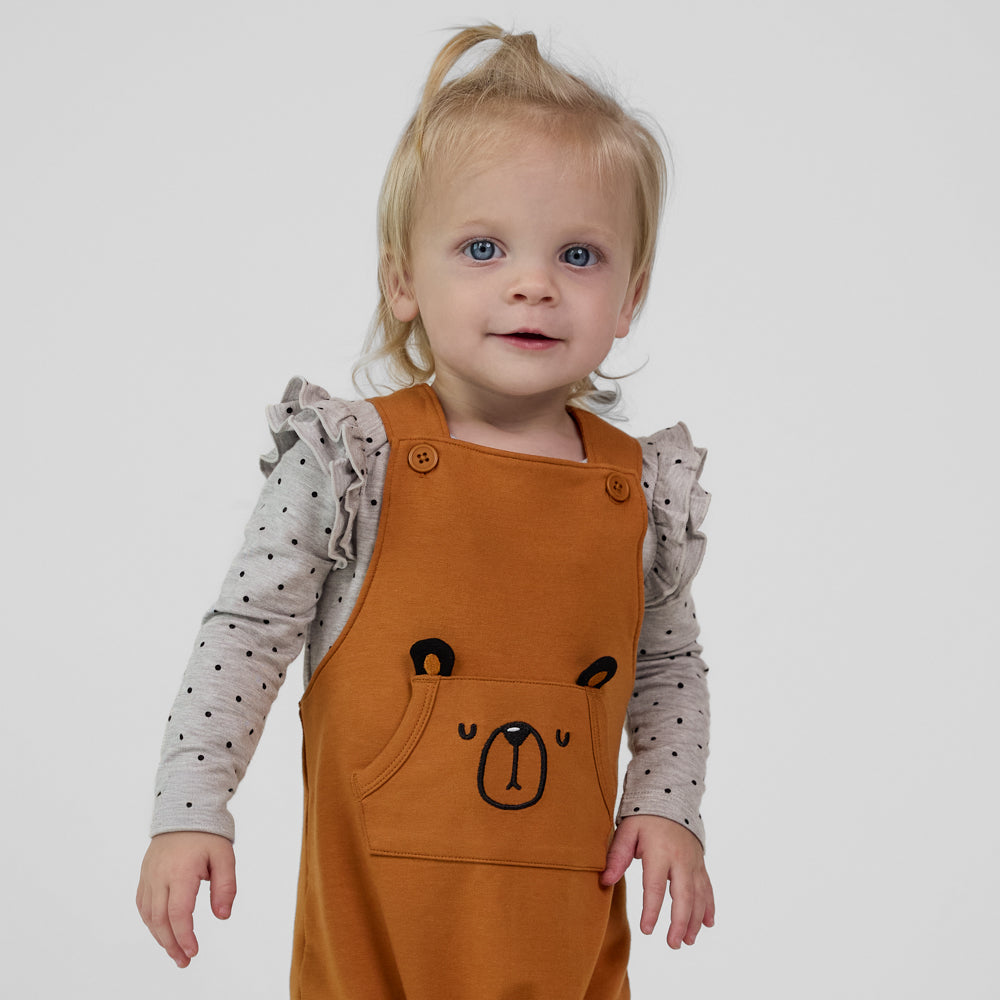 Close-up view of a child wearing a Golden Brown graphic overall paired with a gray long sleeve flutter bodysuit