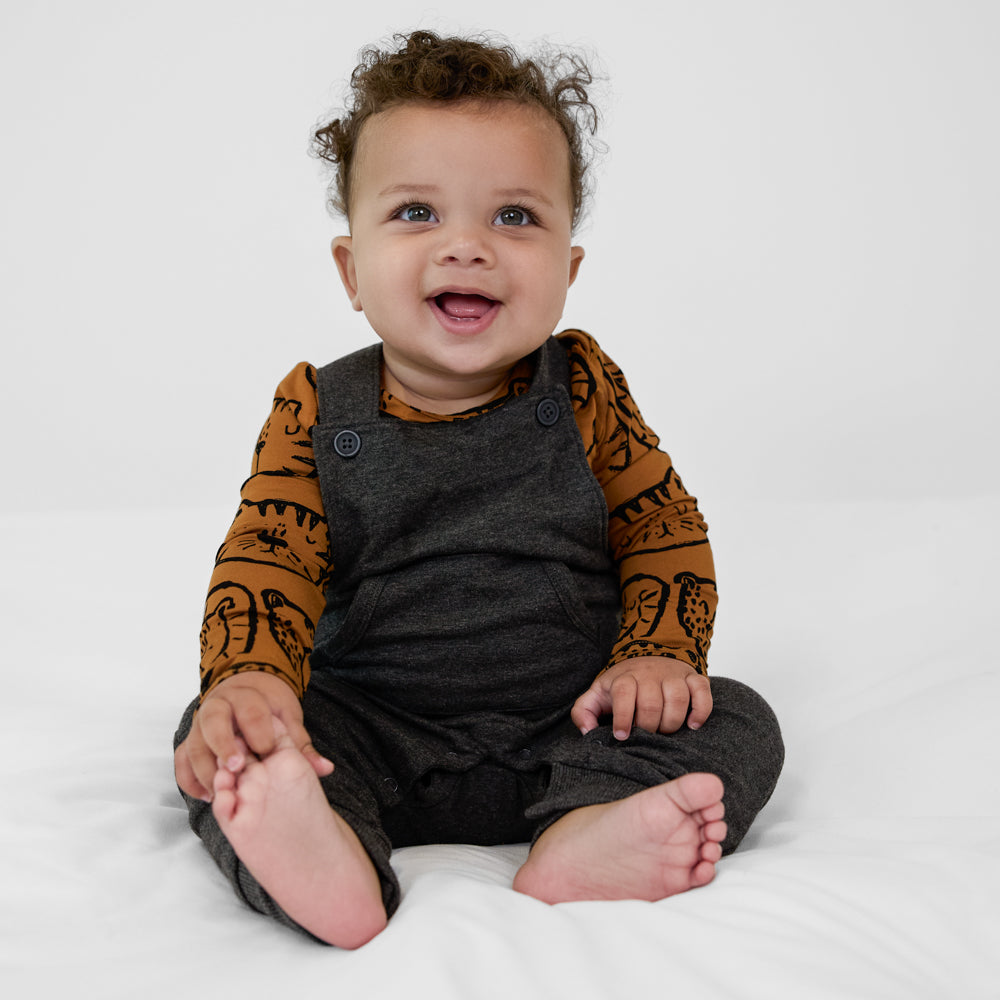 Child sitting on the ground wearing a Heather Black overall paired with an amber with black animal print long sleeve bodysuit