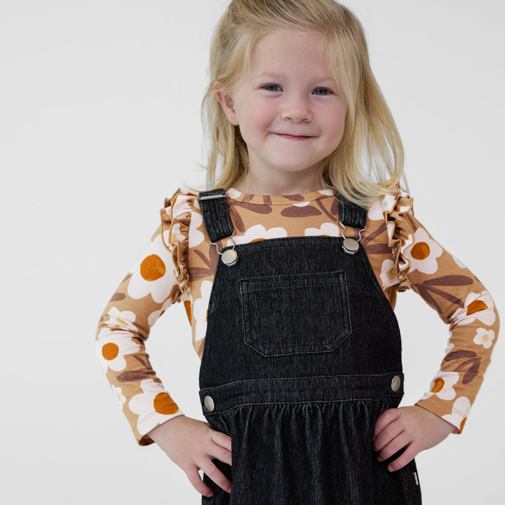 Child wearing a Black denim tank skirt overall paired with a tan floral ruffle shoulder long sleeve top