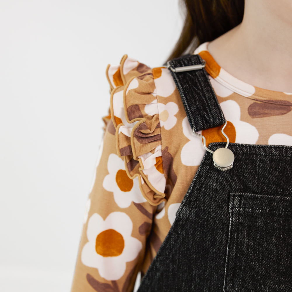 Close-up view of the shoulder of a child wearing a Black denim tank skirt overall with a tan floral ruffle shoulder top