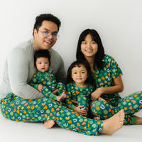 Family of four sitting together wearing matching Lucky printed pajamas. Dad is wearing a men's solid Heather Gray pajama top and Lucky printed men's pajama pants. Mom is wearing a women's Lucky printed pajama top and matching pajama pants. Their children are matching wearing Lucky printed two piece pajama set and zippy.