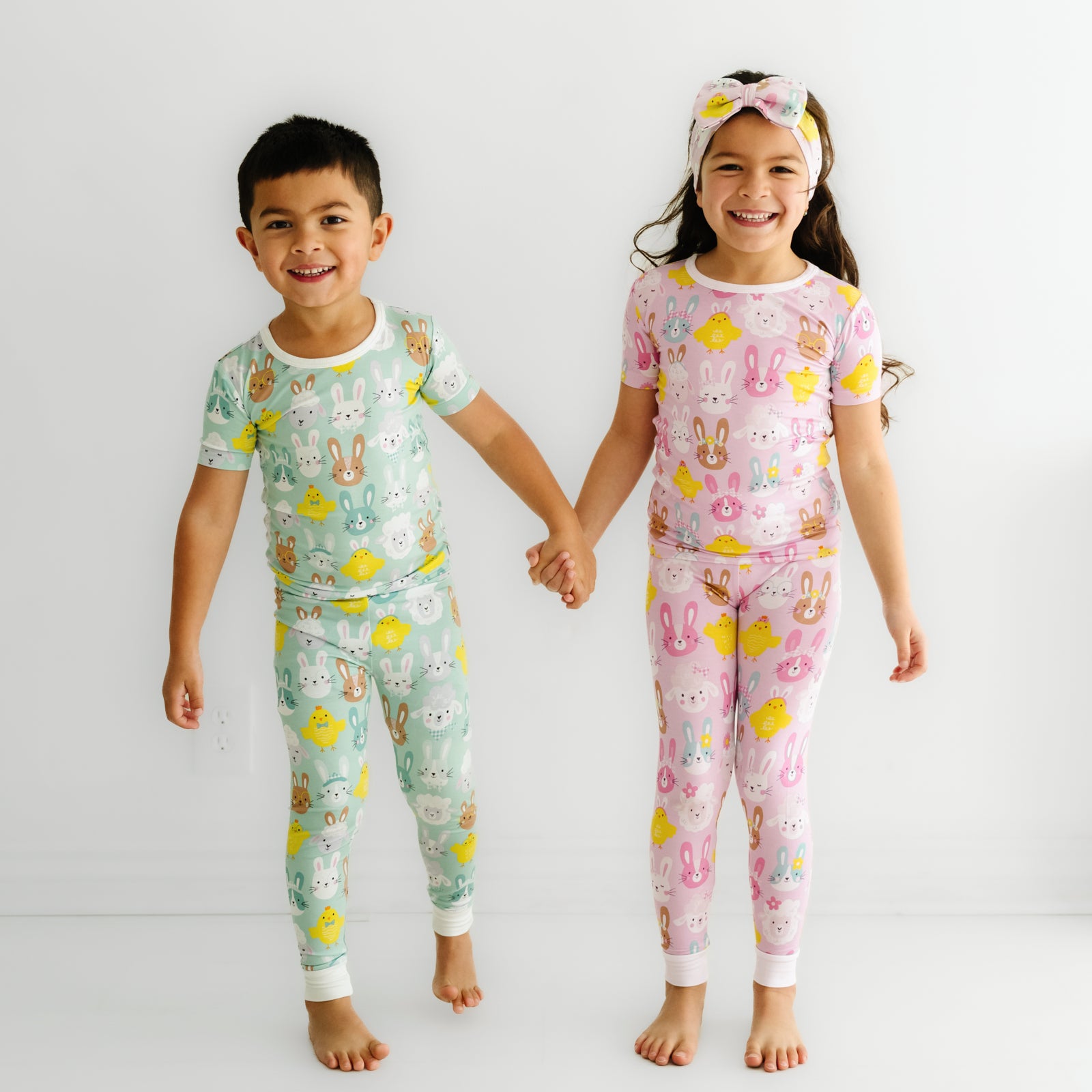 Two children holding hands wearing coordinating Aqua and Pink Pastel Parade two piece short sleeve pajama sets. One is pairing her pajamas with a matching Pink Pastel Parade luxe bow headband