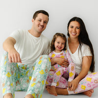 Family of three sitting together. Dad is wearing men's Aqua Pastel Parade pajama pants paired with a men's Bright White short sleeve pajama top. Mom is wearing Pink Pastel Parade women's pajama pants paired with a coordinating Bright White women's pocket tee. Her daughters is matching wearing a Pink Pastel Parade two piece pajama set