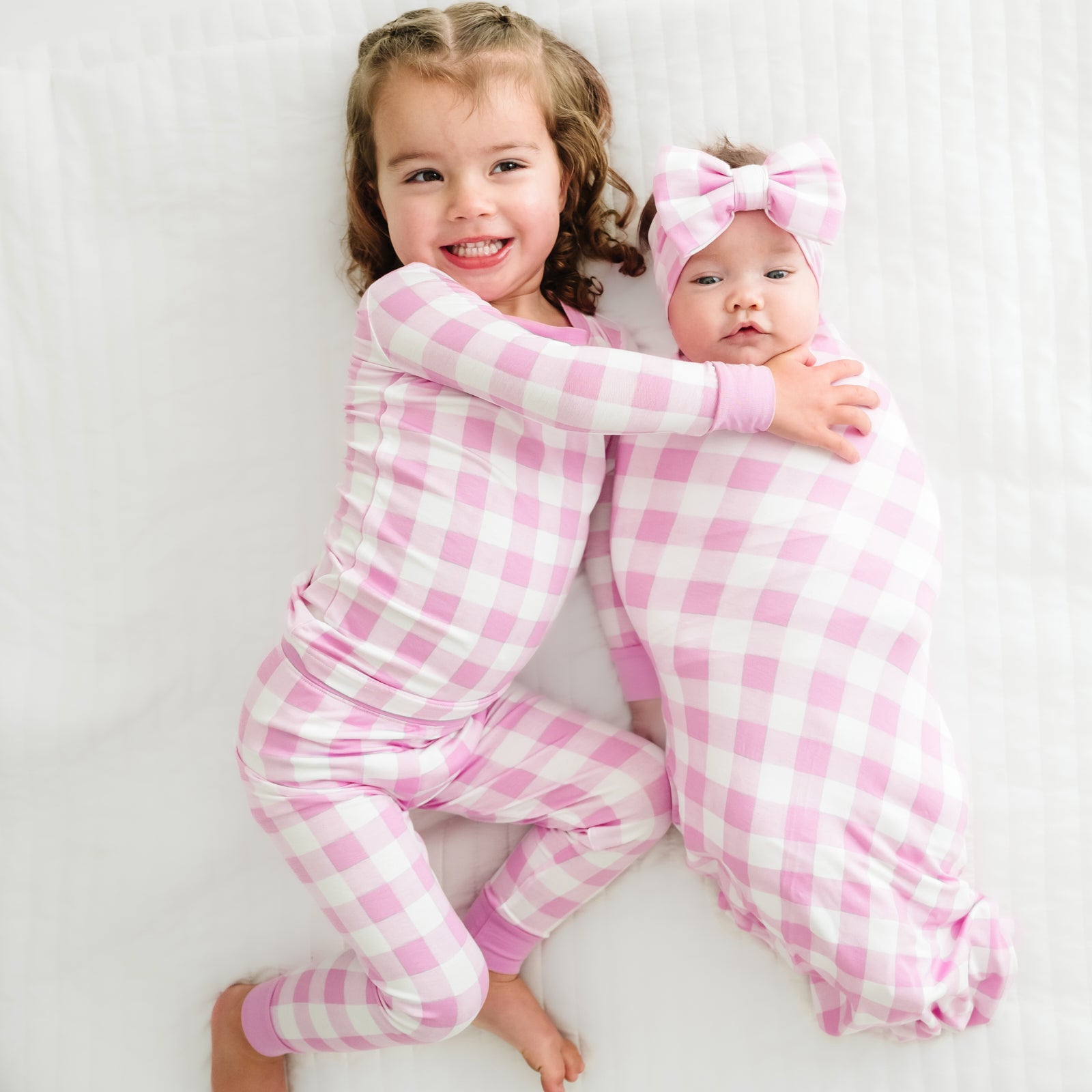 two children cuddling wearing matching Pink Gingham two piece pajama set and child swaddled in a Pink Gingham swaddle and luxe bow headband set