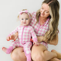 Woman holding her child wearing matching Pink Gingham pajamas. Mom is wearing women's Pink Gingham pajama shorts and matching women's short sleeve pajama top. Child is wearing a matching Pink Gingham zippy paired with a matching luxe bow headband
