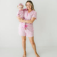 Woman holding her child wearing matching Pink Gingham pajamas. Mom is wearing women's Pink Gingham pajama shorts and matching women's short sleeve pajama top. Child is wearing a matching Pink Gingham zippy paired with a matching luxe bow headband 