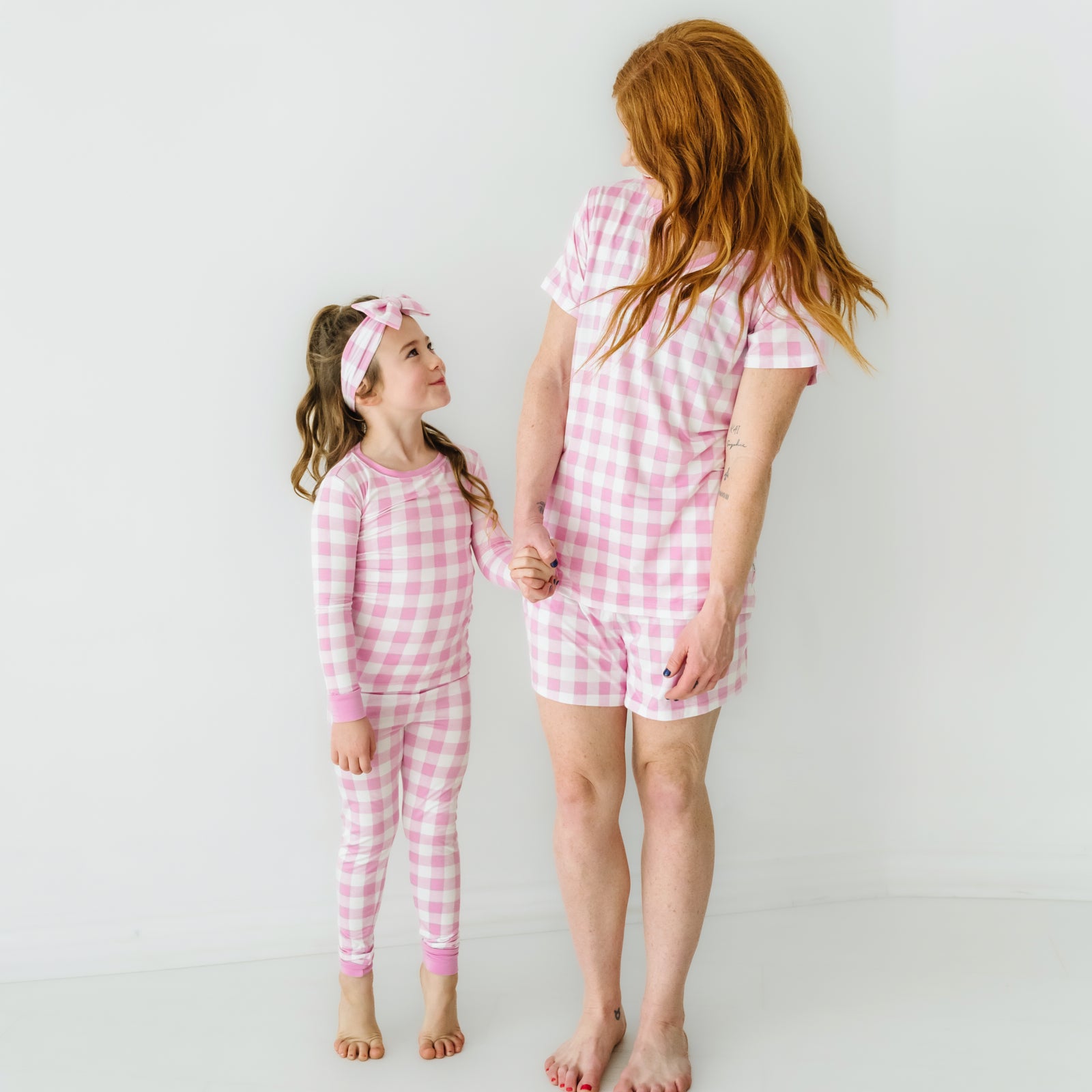 Mother and daughter matching wearing Pink Gingham. Child is wearing a Pink Gingham two piece pajama set paired with a matching luxe bow. Mom is wearing a Pink Gingham women's short sleeve pajama top and matching women's pajama shorts