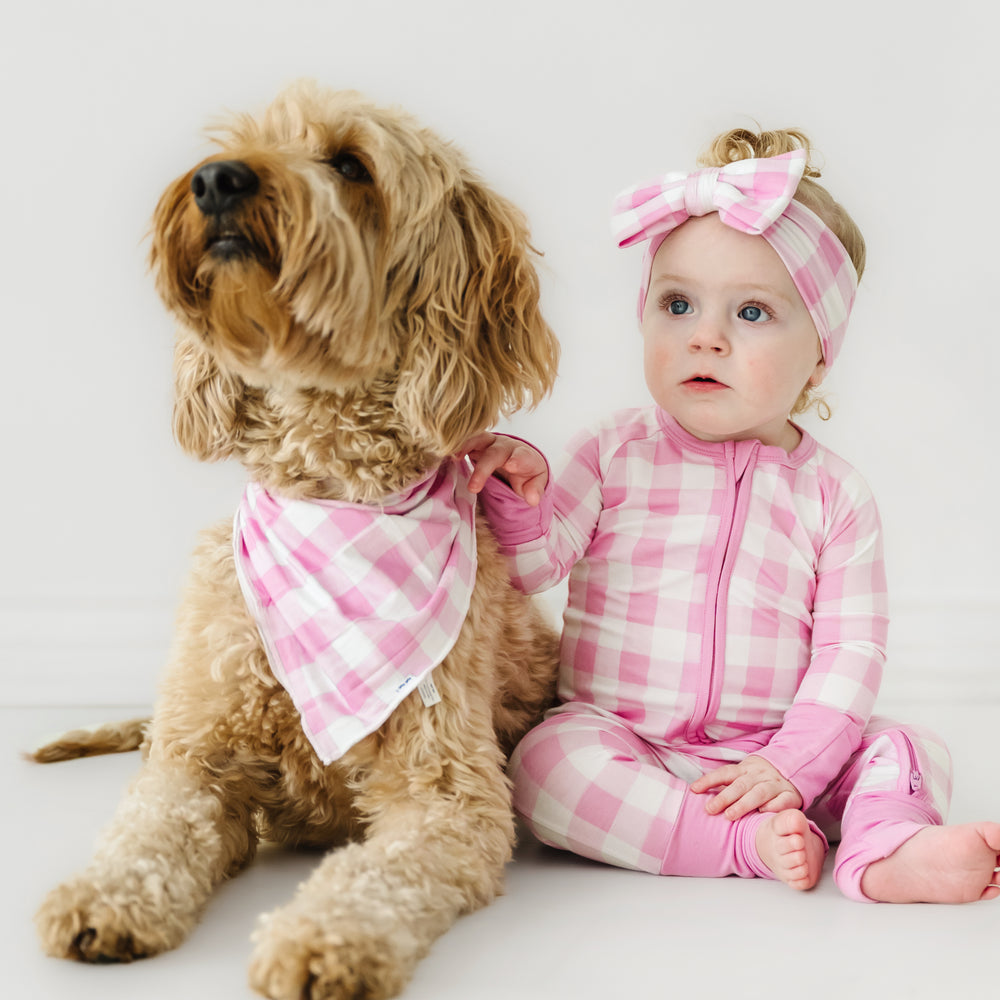 Click to see full screen - Child sitting next to her dog wearing matching Pink Gingham. Child is wearing a Pink Gingham zippy paired with a matching luxe bow. Her dog is wearing a Pink Gingham pet bandana 