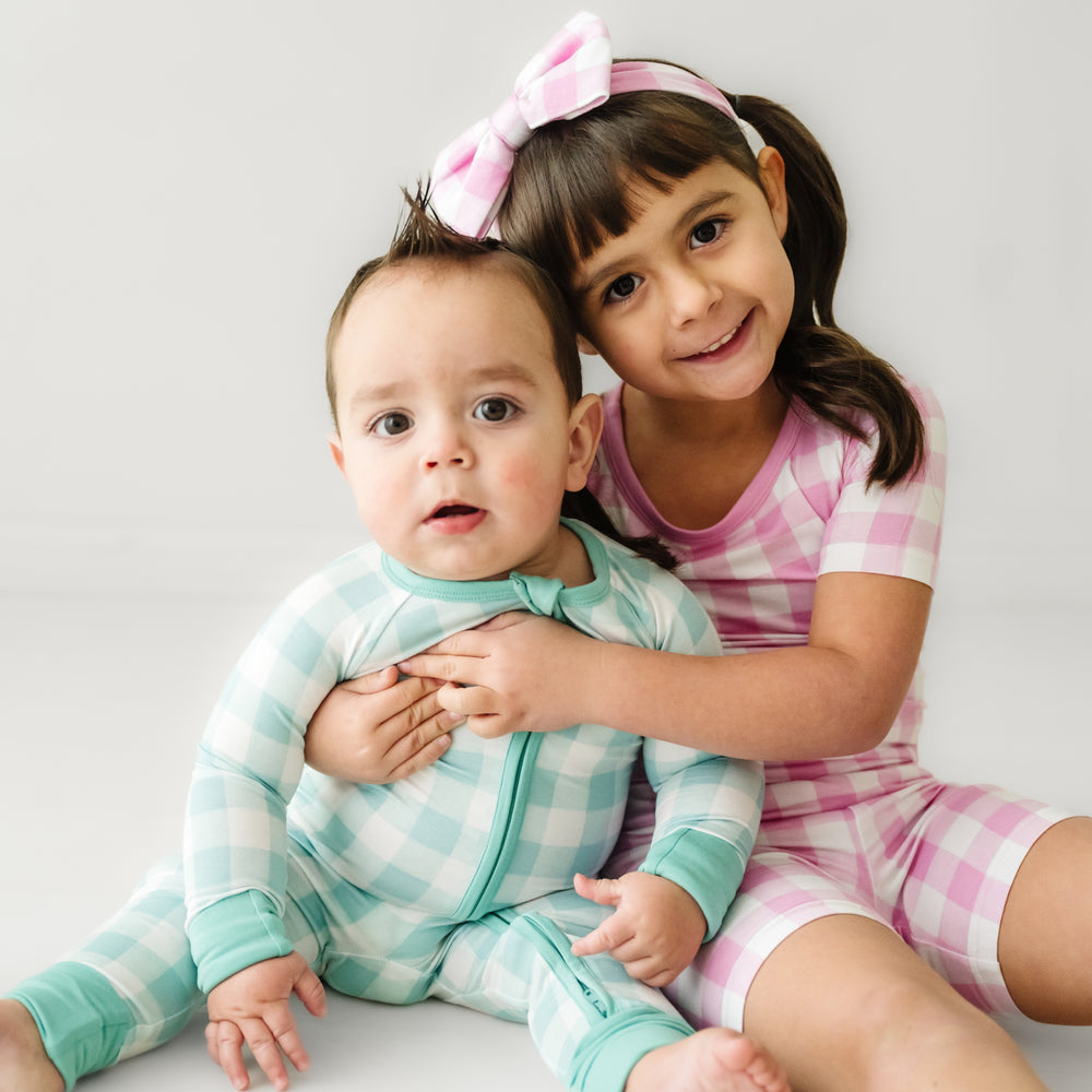 Click to see full screen - Two children snuggling. One child is wearing a Pink Gingham two piece short sleeve and shorts pajama set paired with a matching luxe bow headband. Her brother is matching wearing an Aqua Gingham zippy