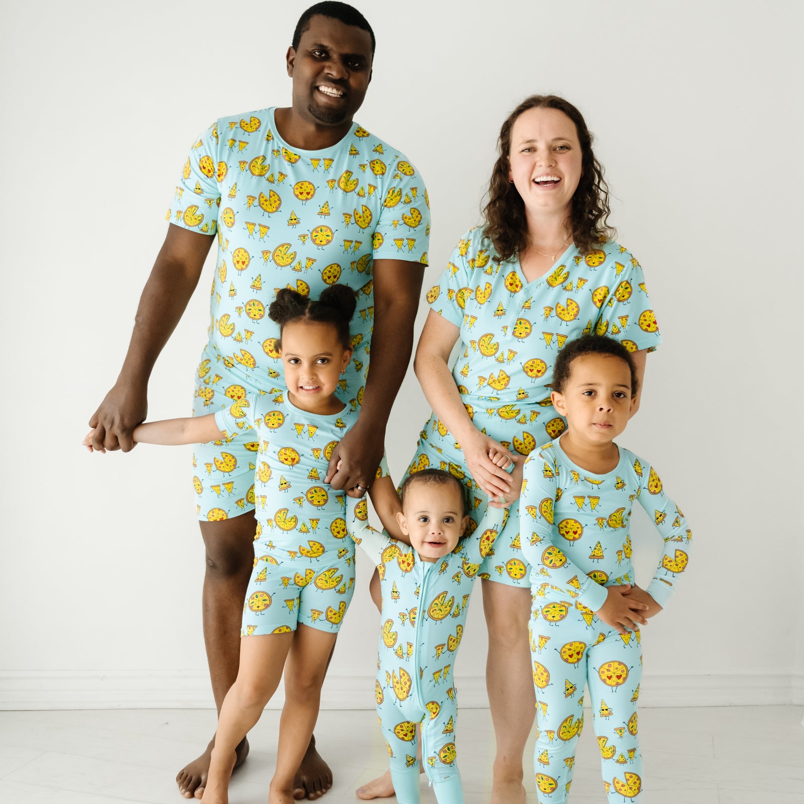 Family of five wearing matching pjs. Mom is wearing a women's Pizza Pal pajama top and matching women's pajama shorts. Dad is wearing men's Pizza Pals men's pj top and matching men's pj shorts. Kids are wearing matching Pizza Pals pjs in two piece and zippy styles