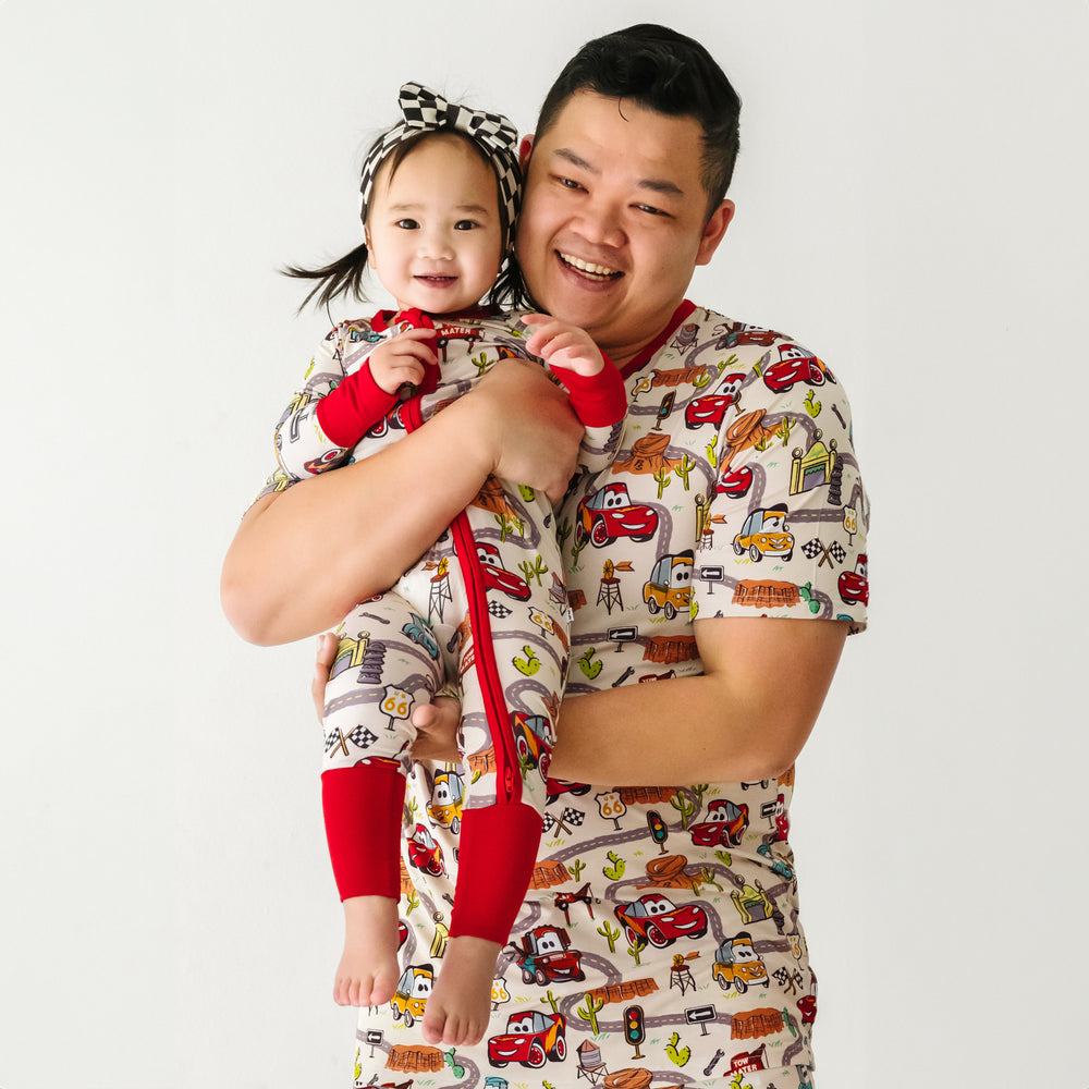 Close up image of a dad holding his daughter wearing matching Radiator Springs pjs. Dad is wearing men's Radiator Springs pj top, daughter is wearing matching Radiator Springs zippy and a racing checks luxe bow