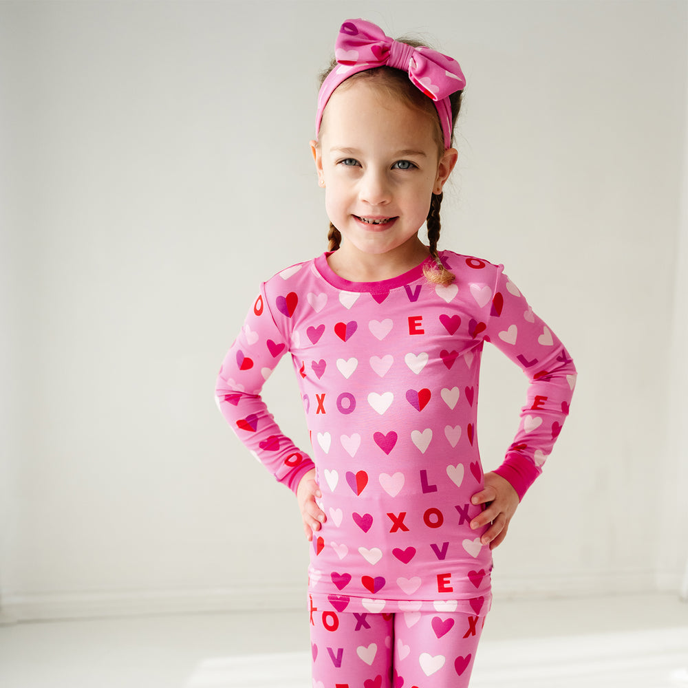 Click to see full screen - Close up image of a child posing wearing Pink XOXO two piece pajama set paired with a matching luxe bow headband