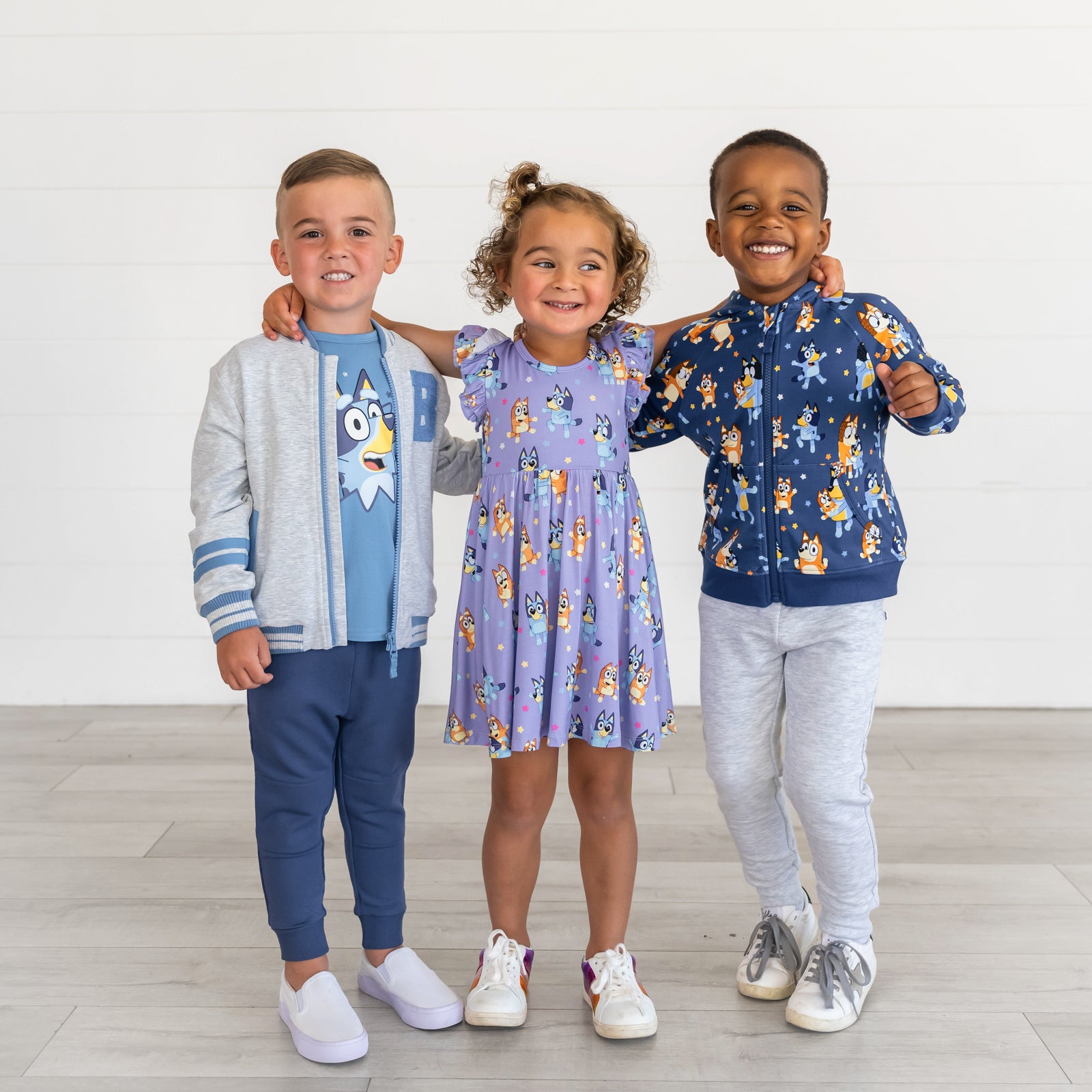 Boy on the left is wearing the Gray Bomber Jacket & Bluey Graphic Tee. Girl in the middle Bluey & Bingo Flutter Twirl Dress. Boy on the right is wearing the Dance Mode Zip Hoodie