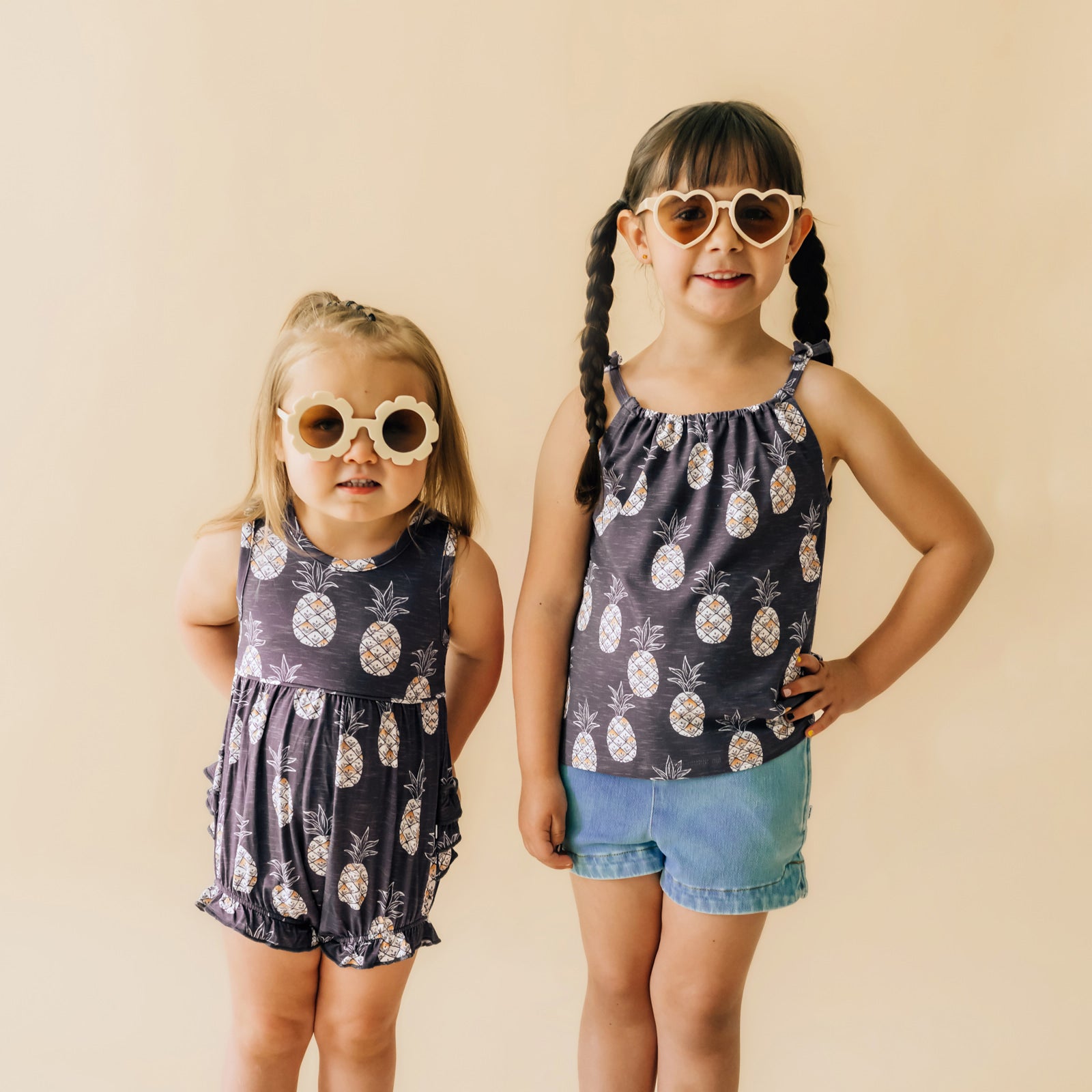 Two children posing together wearing coordinating Sweet Paradise play items. One child is wearing a Sweet Paradise bubble romper and the other child is wearing a Sweet Paradise flowy bow strap tank paired with light blue denim shorts