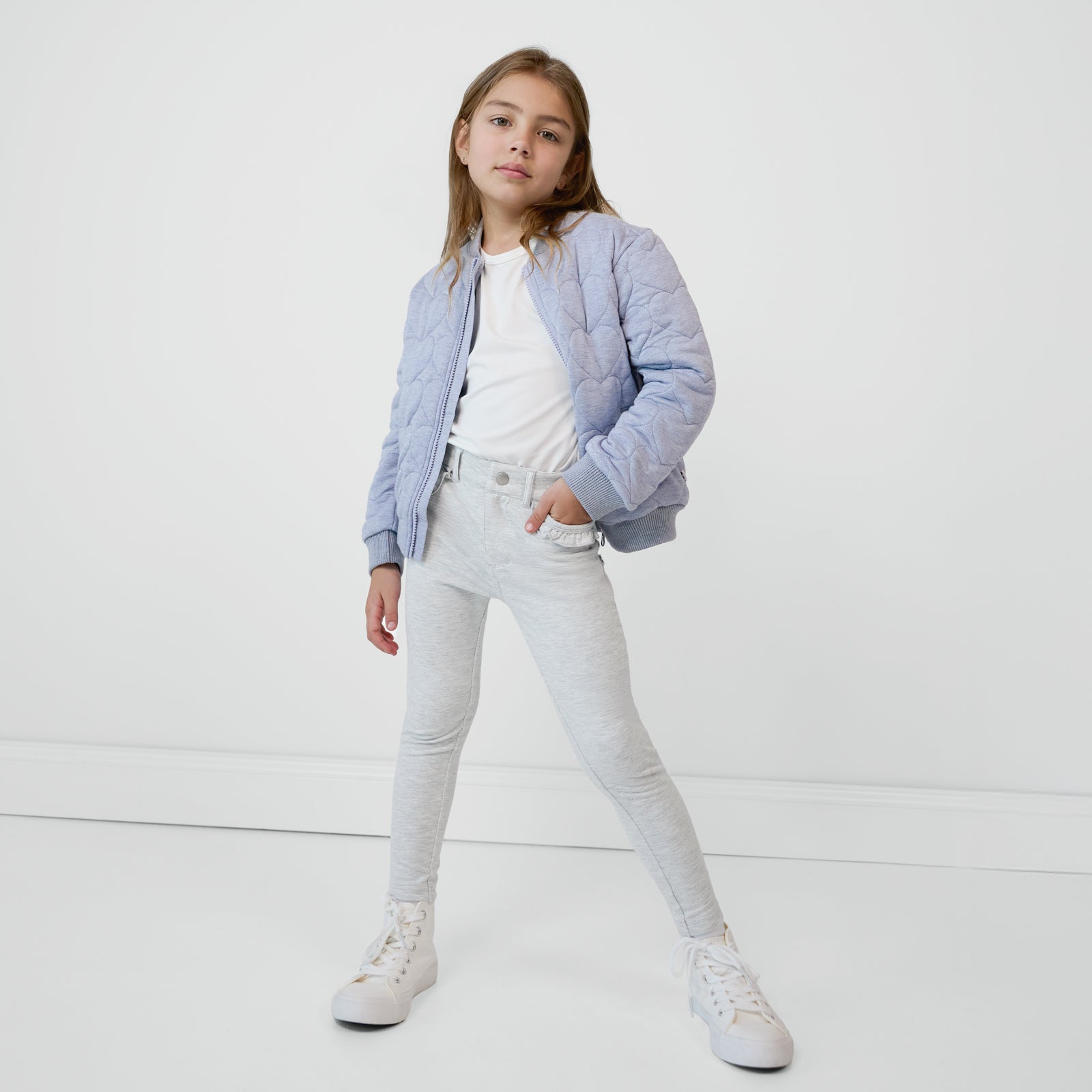 Child with their hand in a pocket wearing Light Heather Gray Ruffle Heart Pocket Pants paired with a soft white top and quilted jacket