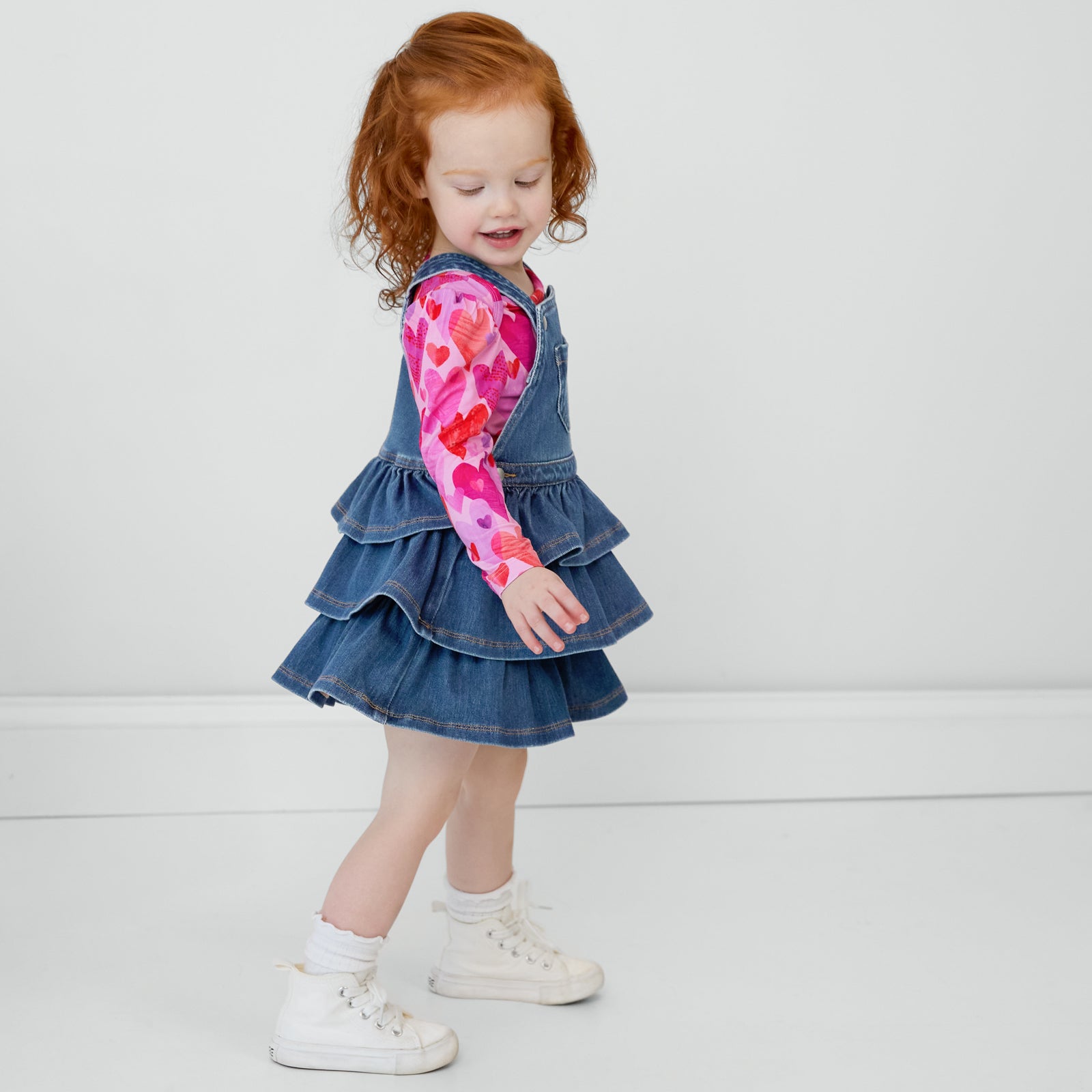 Side view of a child wearing a Midwash Blue Denim Tiered Skirt Overall and coordinating Hearts & Crafts top underneath