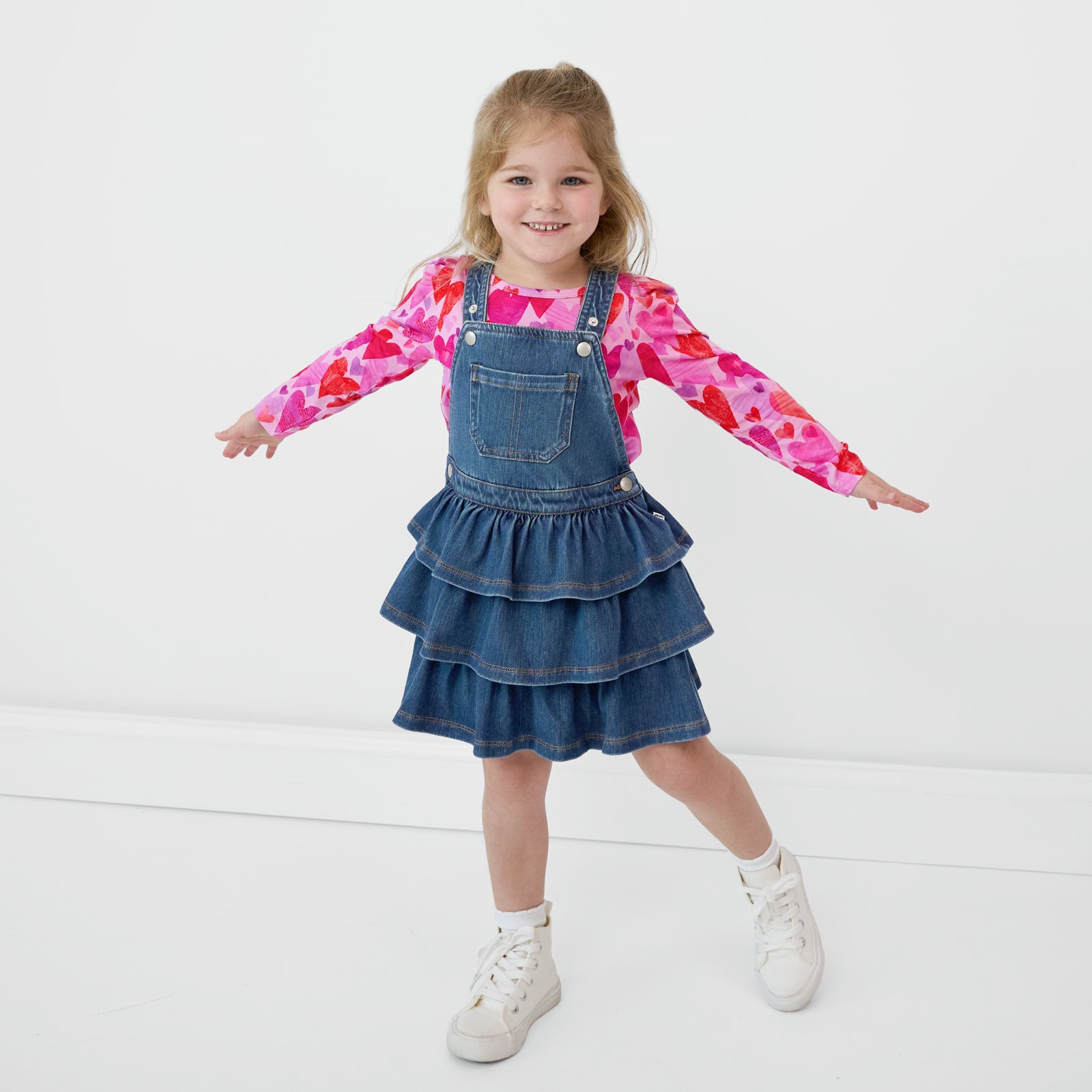 Smiling child with her arms out wearing a Midwash Blue Denim Tiered Skirt Overall and a coordinating Hearts & Crafts top underneath