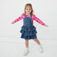 Smiling child with her arms out wearing a Midwash Blue Denim Tiered Skirt Overall and a coordinating Hearts & Crafts top underneath