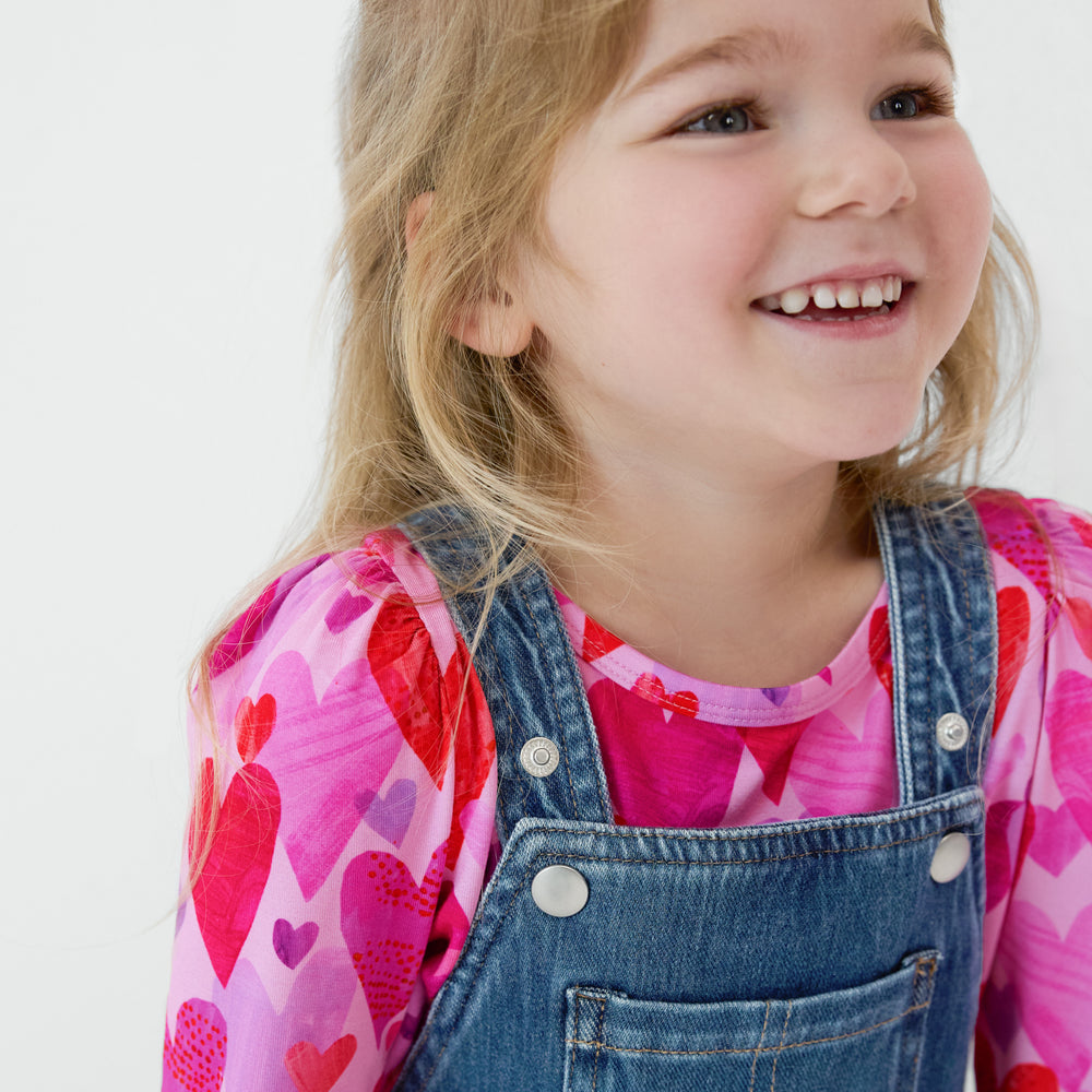 Close up of a smiling child wearing a Midwash Blue Denim Tiered Skirt Overall with a coordinating Hearts & Crafts top underneath