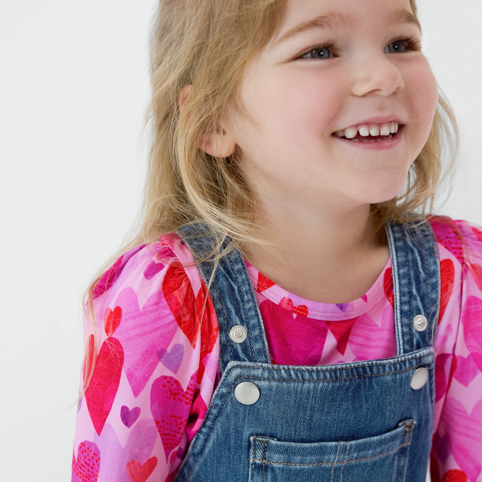 Close up of a smiling child wearing a Midwash Blue Denim Tiered Skirt Overall with a coordinating Hearts & Crafts top underneath