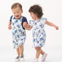 Two children playing wearing matching Seas the Day play items. One child is wearing a Seas the Day shorty overall paired with a vintage navy bodysuit and the other child is wearing a Seas the Day printed flutter skater dress with bodysuit