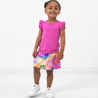 child posing wearing a Rouge Pink Flutter Tee paired with a Rainbow Blooms skort