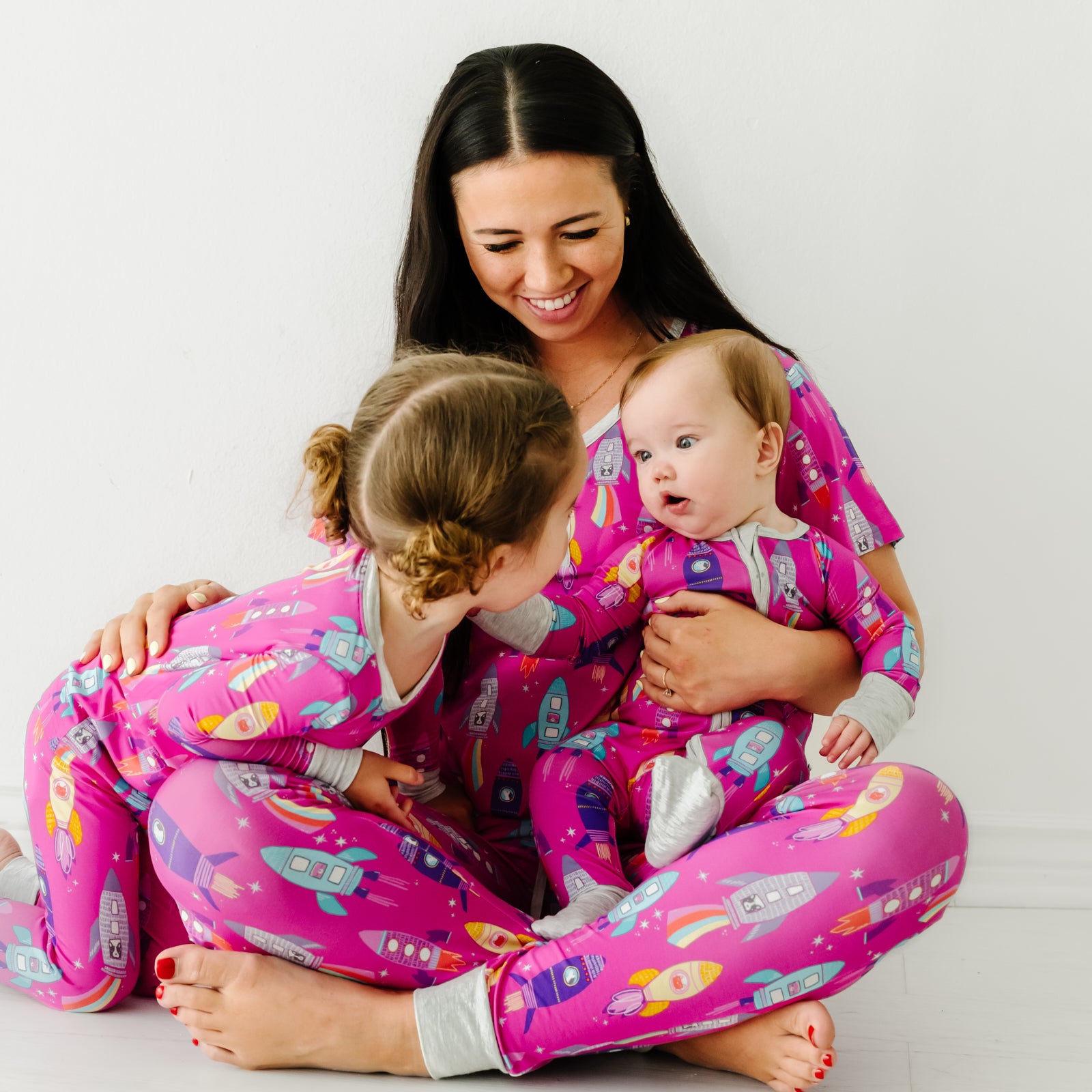 Mother sitting with her two children wearing matching Pink Space Explorer pajama sets. Mom is wearing a women's Pink Space Explorer pajama top and matching pajama pants. Her children are wearing Pink Space Explorer pajamas in two piece and zippy styles