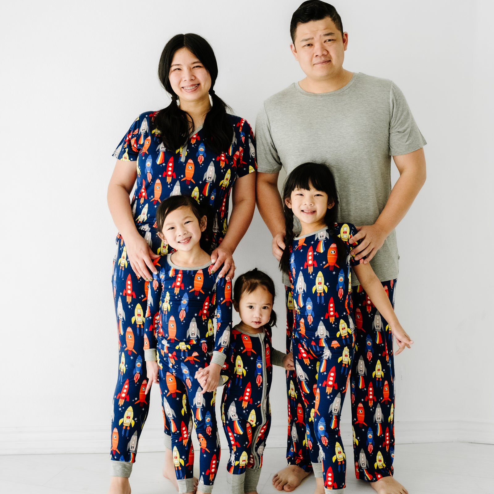 Family of four posing together wearing matching Navy Space Explorer pajama sets. Mom is wearing women's Navy Space Explorer women's pajama top paired with matching women's pajama pants. Dad is wearing a solid heather gray men's pajama top paired with men's Navy Space Explorer pajama pants. Their three children are wearing Navy Space Explorer pajama sets in two piece and zippy styles