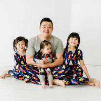 Father sitting with his three children matching wearing Navy Space Explorer pajamas. Dad is wearing men's Navy Space explorer pajama pants paired with a Heather Gray men's pajama top. His kids are wearing Navy Space Explorer pajamas in two piece and zippy styles