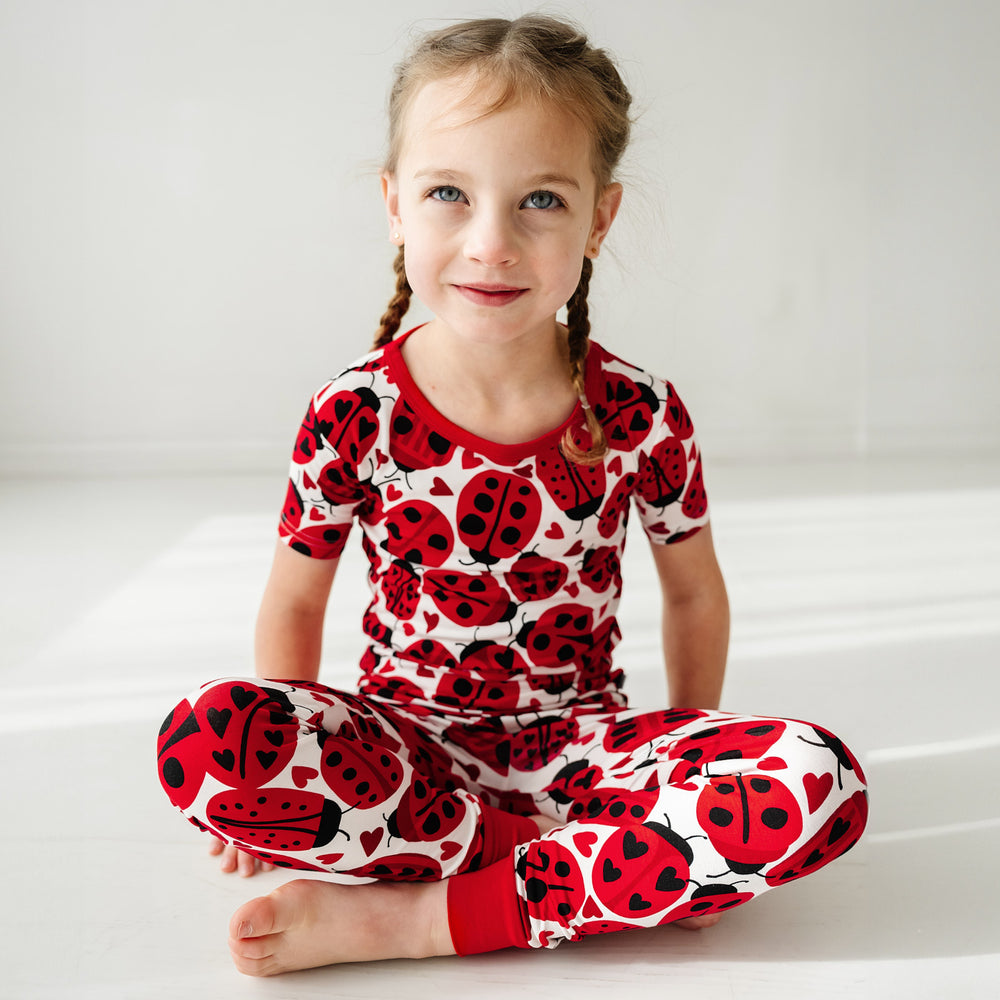 Click to see full screen - Child sitting on the ground wearing a Love Bug printed two-piece short sleeve pajama set