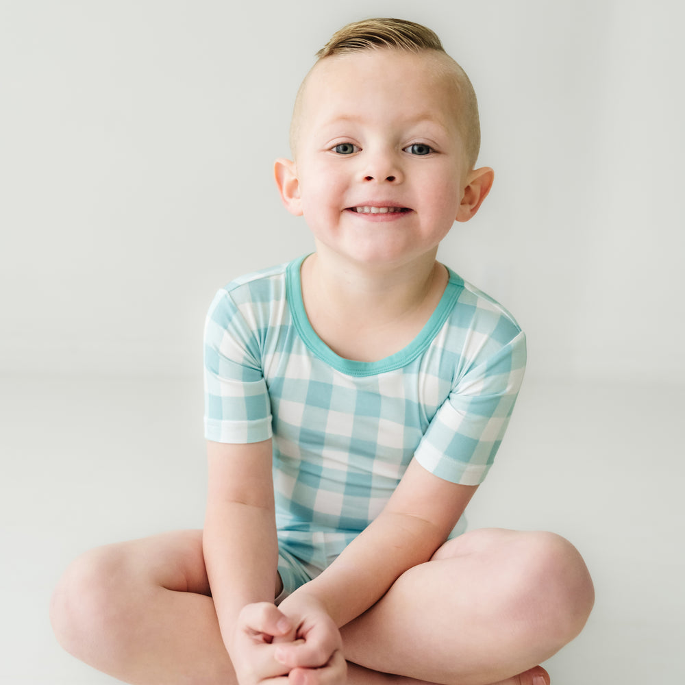 Click to see full screen - Child sitting wearing a Aqua Gingham two piece short sleeve and shorts pajama set
