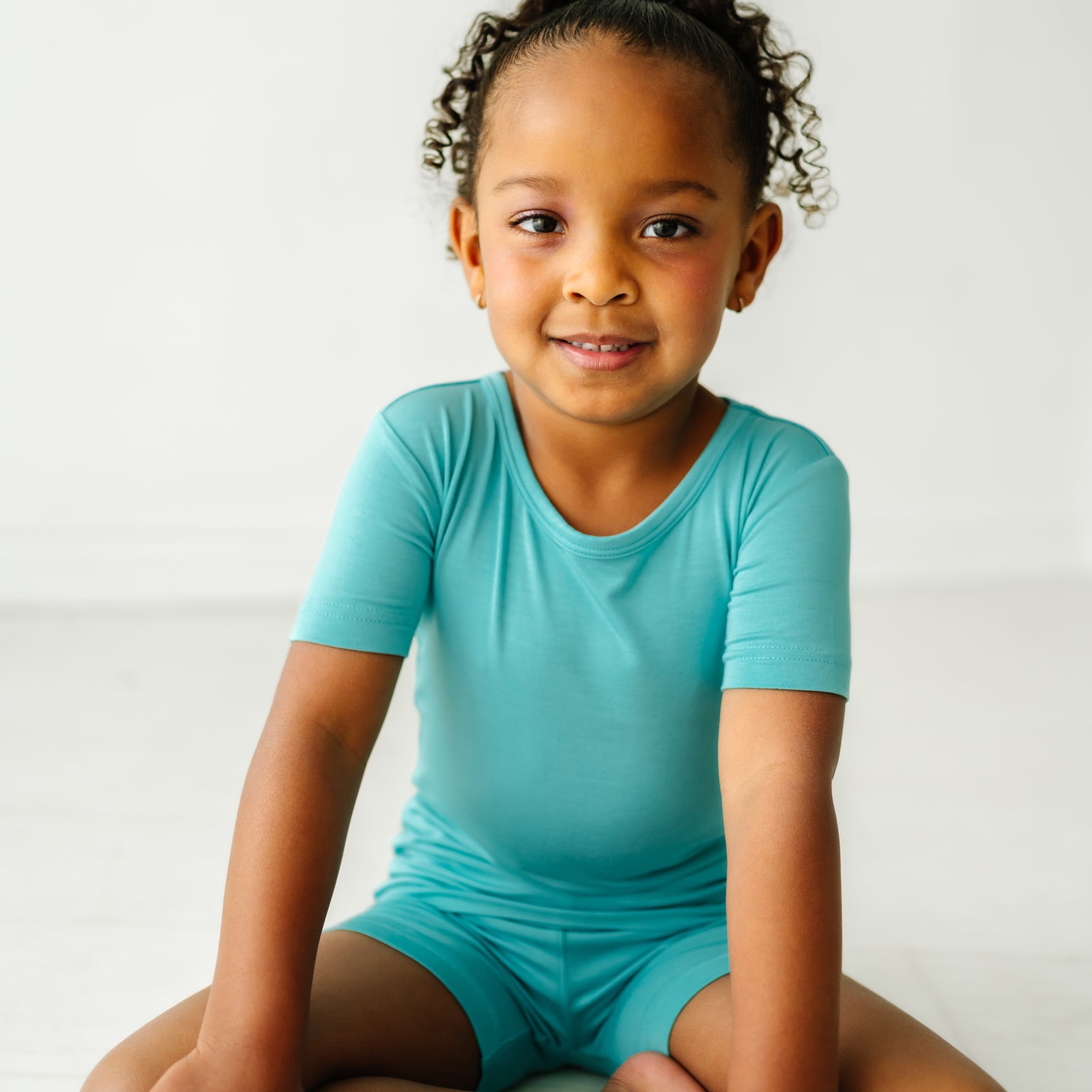 Close up image of a child sitting wearing a Glacier Turquoise two piece short sleeve and shorts pajama set