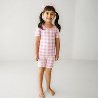 Child wearing a Pink Gingham two piece short sleeve and shorts pajama set paired with a matching luxe bow headband